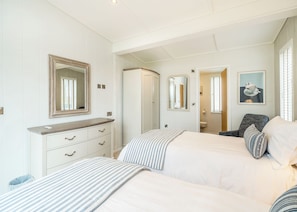 Une chambre à coucher avec un lit, une commode, un miroir, une chaise et une salle de bain visible à travers une porte ouverte.