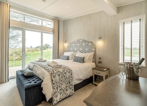 A bedroom with a large bed, a bench, a nightstand, a bedside table with a bottle and glasses, and a view of the outdoors.