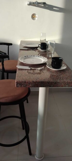 A dining area with a table for two, featuring a white plate, black mug, and clear glass, set amidst a room with a tiled floor and a white wall.
