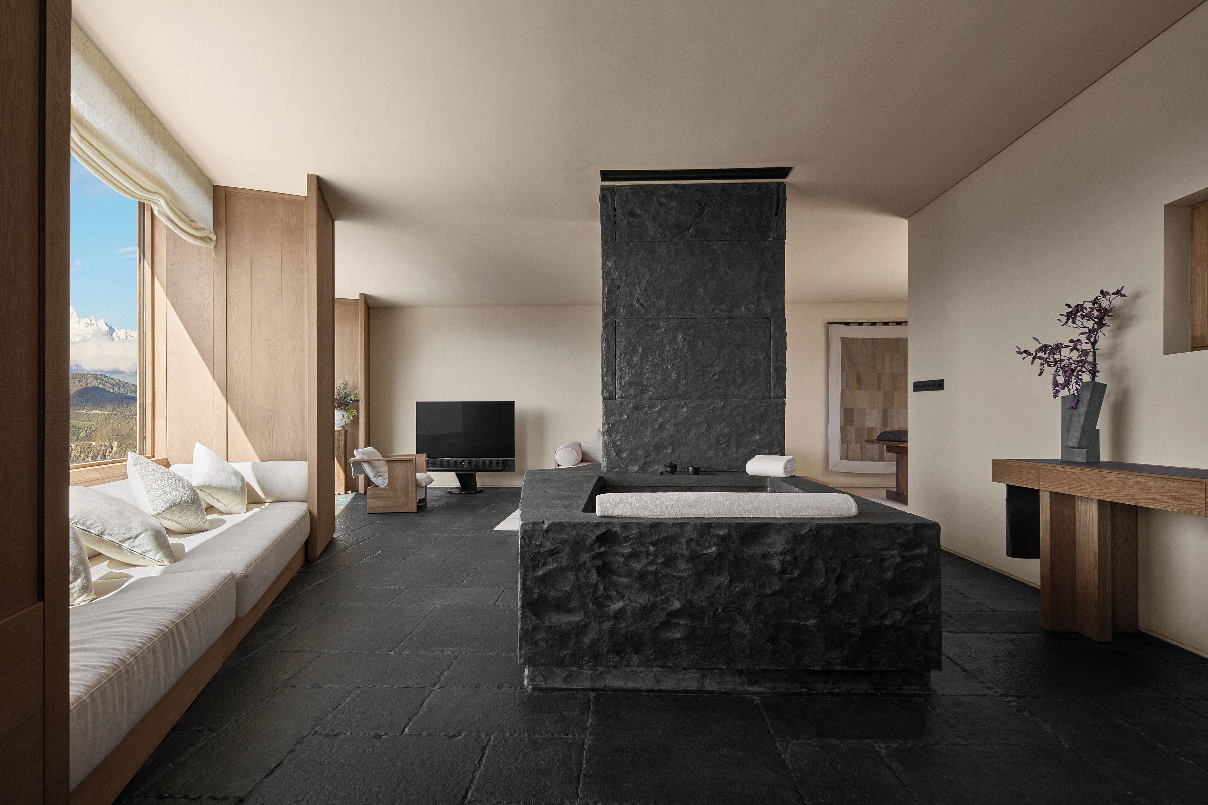 Een moderne badkamer met een stenen wastafel, een houten bankje en een groot raam met uitzicht op een bergachtig landschap.