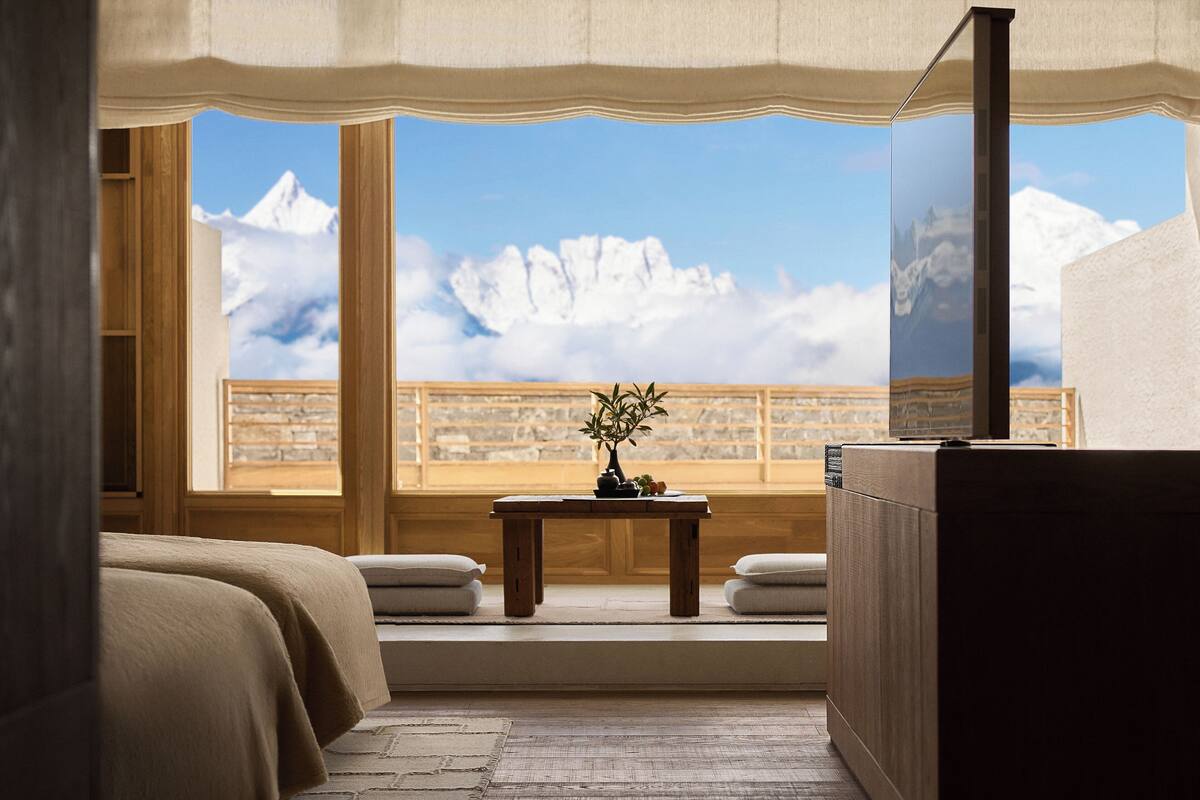 Une chambre avec une grande fenêtre, une table en bois ornée d’un vase et un lit recouvert de draps blancs.