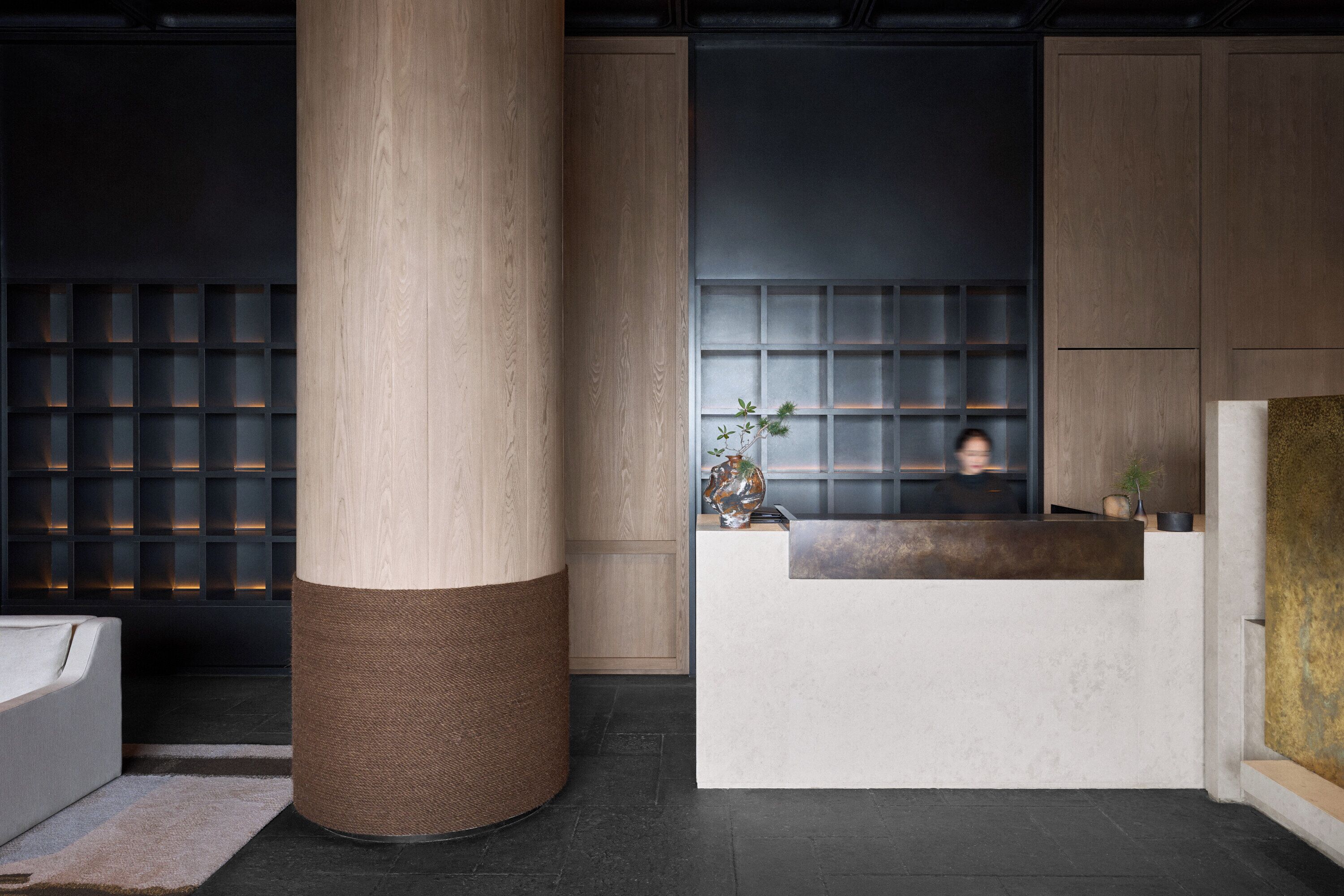 Zona de recepción con un mostrador de madera, un pilar grande y una pared con diseño de cuadrícula.