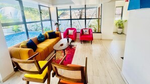 Ein modernes Wohnzimmer mit einem gelben Sofa, roten Sesseln und einem Couchtisch aus Holz. Große Fenster bieten Blick auf einen Pool und Grünflächen.