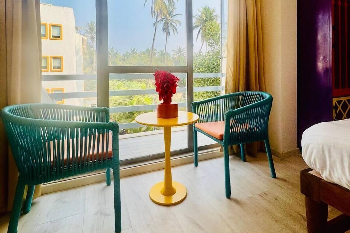 Ein Zimmer mit einem großen Fenster, zwei grünen Stühlen, einem kleinen runden Tisch mit einer Vase und einem Bett.