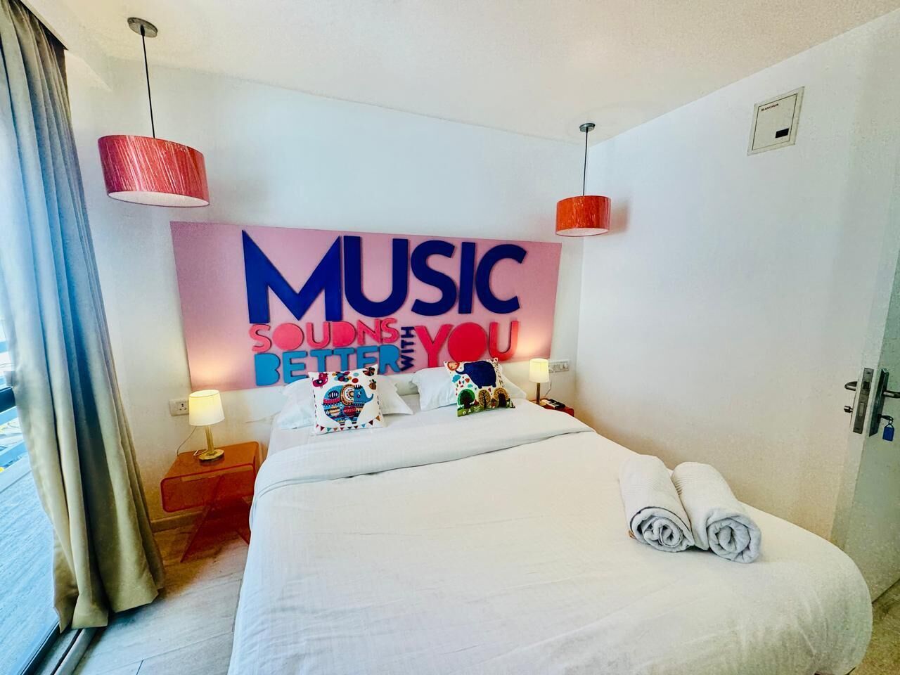A hotel room with a bed, a wall with a 'MUSIC SOUNDS YOU BETTER' sign, a red pendant light, and a window with curtains.
