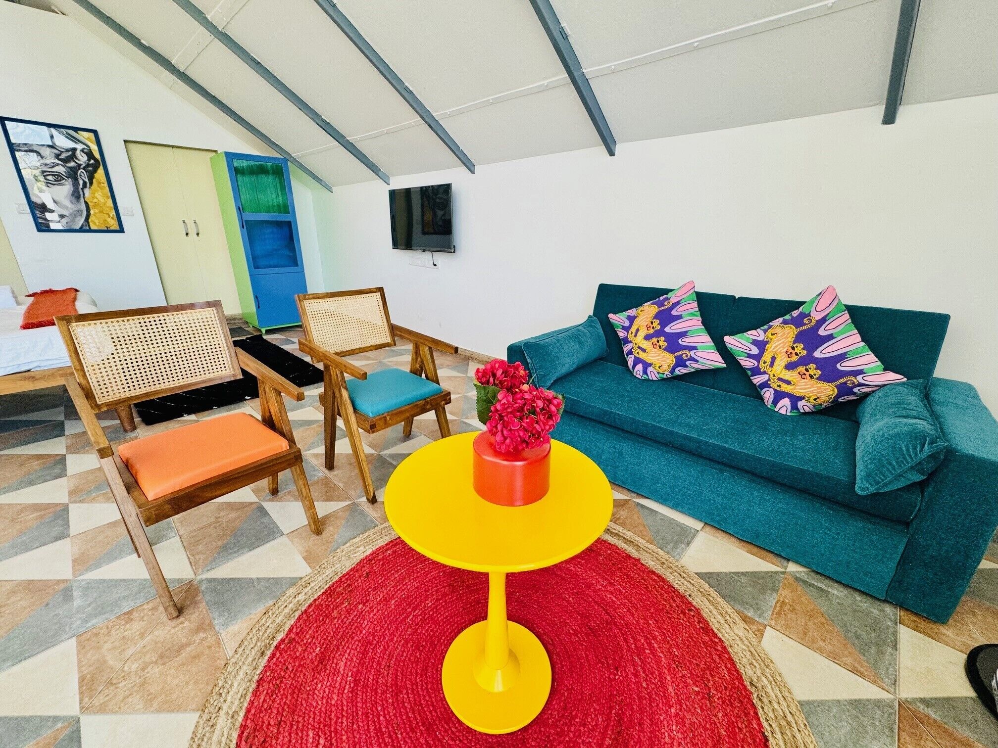 A living room with a teal sofa, colorful cushions, a wooden chair, a yellow table, and a red rug.