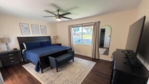 Ein Schlafzimmer mit Bett, Nachttischen, Deckenventilator, großem Fenster mit Vorhängen, Spiegel und Fernseher.