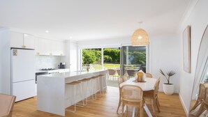 A modern kitchen with white cabinetry, a central island with bar stools, and a dining area with wicker chairs and a white table. - Palm Springs by the Beach, Glam, Pet-Friendly, 4 Bed Retreat incl HUGE studio. (Mollymook Beach)