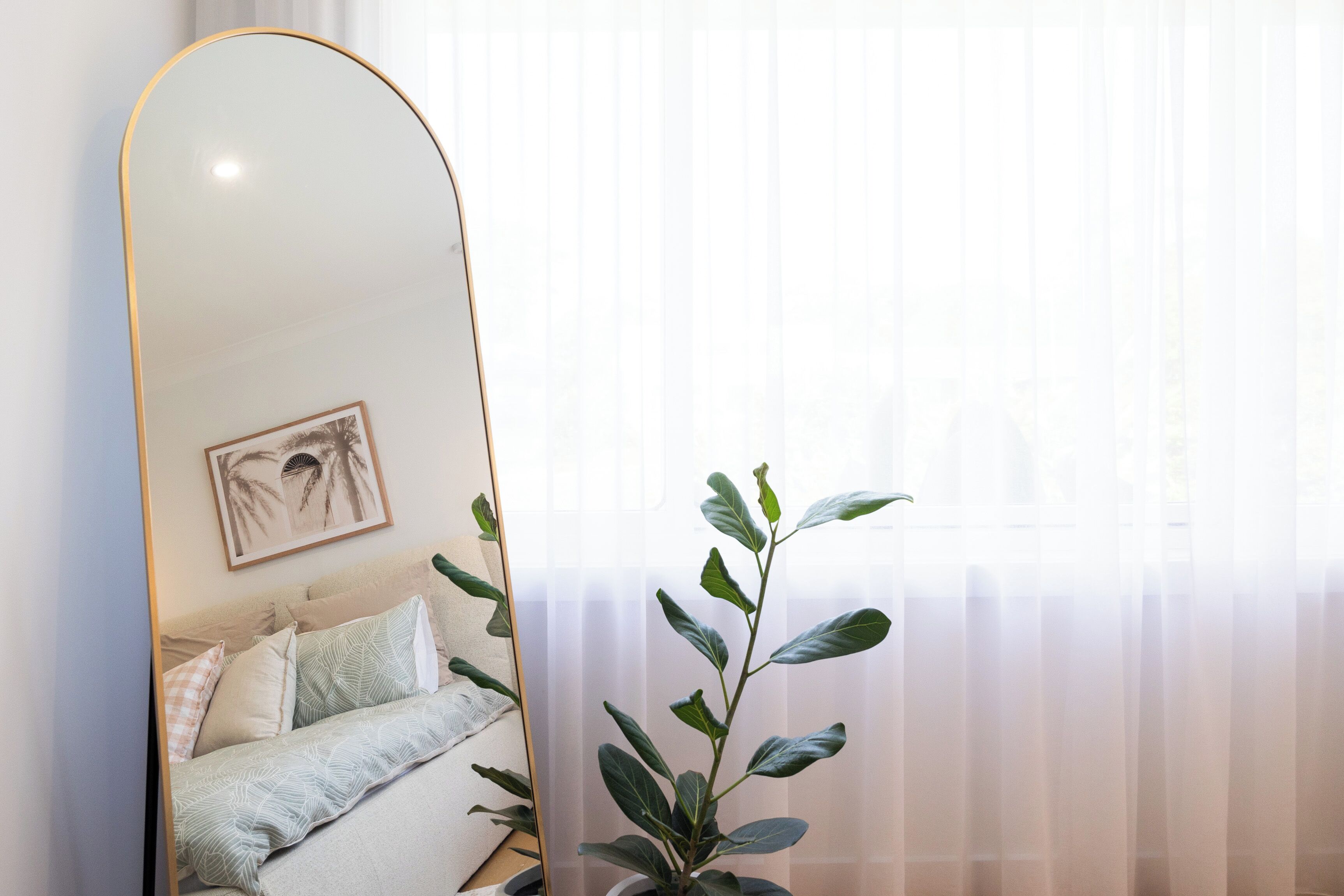 A bedroom with a bed, a bedside plant, a framed picture on the wall, and a window with sheer curtains.