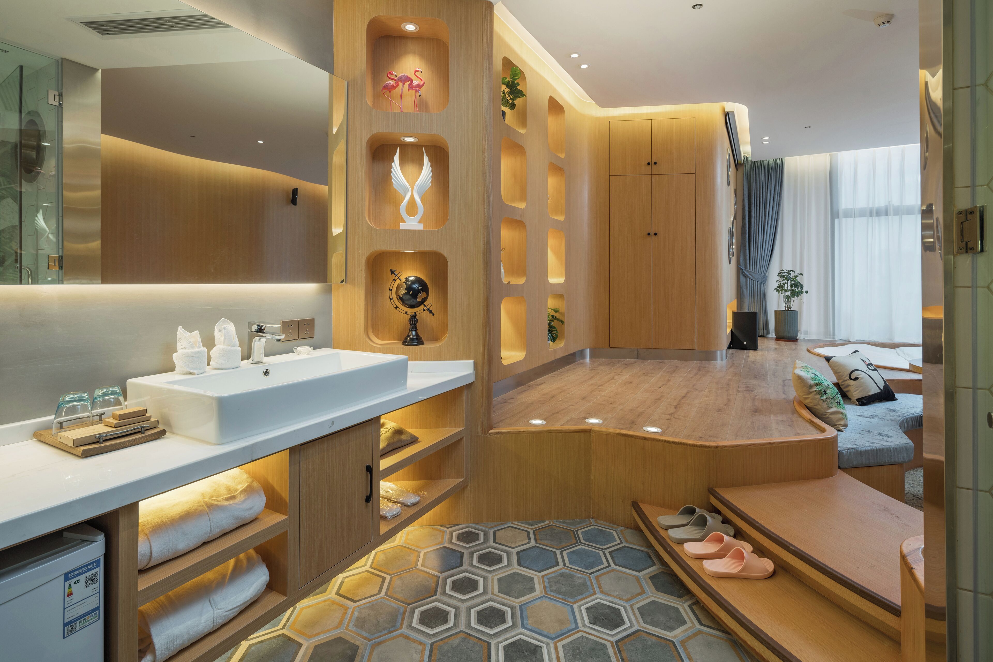 Une chambre d’hôtel moderne avec une coiffeuse en bois, un miroir rond, un lavabo blanc et un sol à motifs hexagonaux.