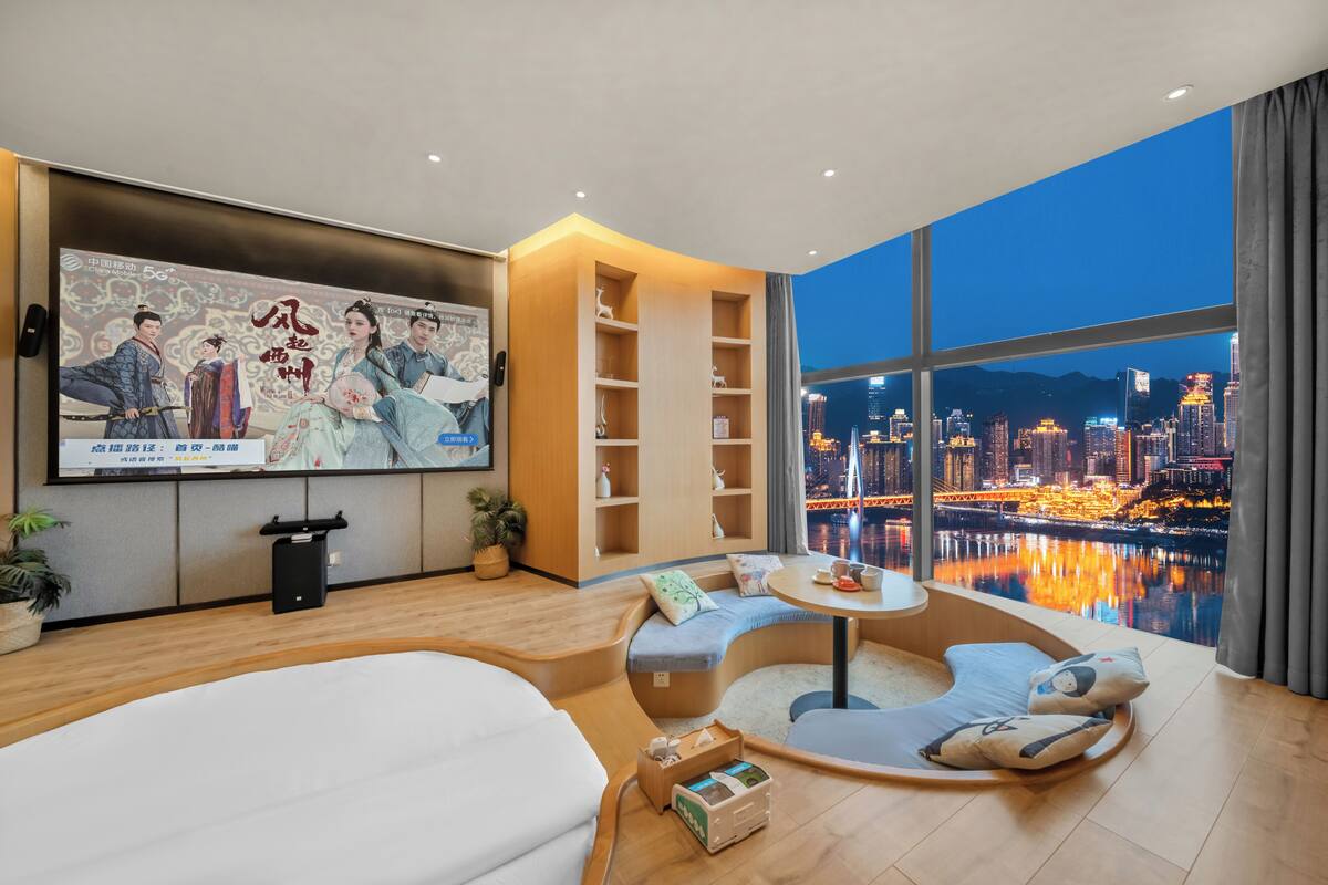 Une chambre d’hôtel moderne dotée d’une grande fenêtre donnant sur la ville, d’un coin salon confortable, d’un téléviseur à écran plat et d’une bibliothèque.