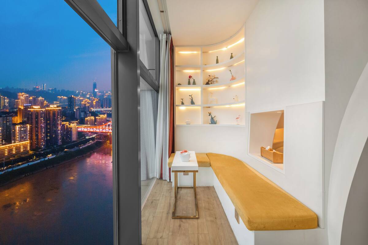 Une pièce moderne avec un banc incurvé, une petite table et des étagères ornées d’objets décoratifs. On peut voir à travers la fenêtre un paysage urbain avec une rivière et des bâtiments illuminés.
