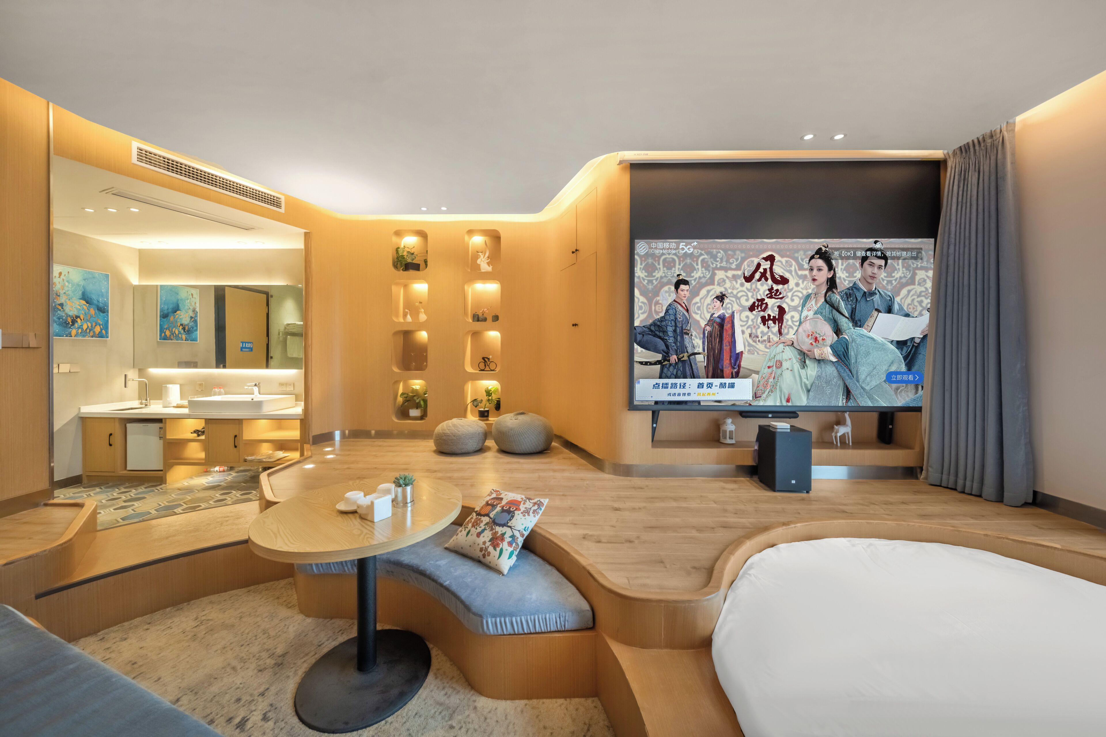 A modern hotel room with a round dining table, a sofa, a TV with a poster, and a bathroom visible through an open door.