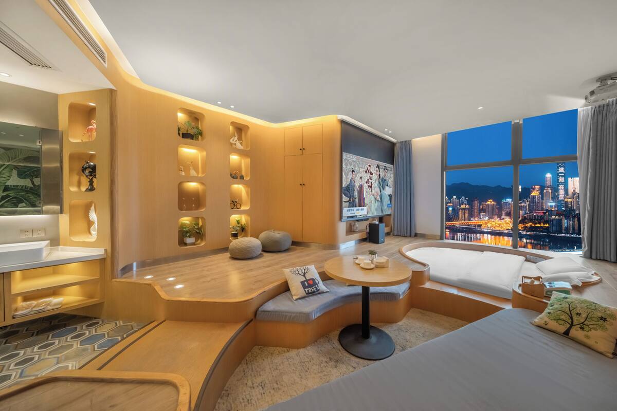 Une chambre d’hôtel moderne dotée d’un espace salon aux lignes courbes, d’une table ronde, d’un lit et d’une grande fenêtre offrant une vue sur la ville.