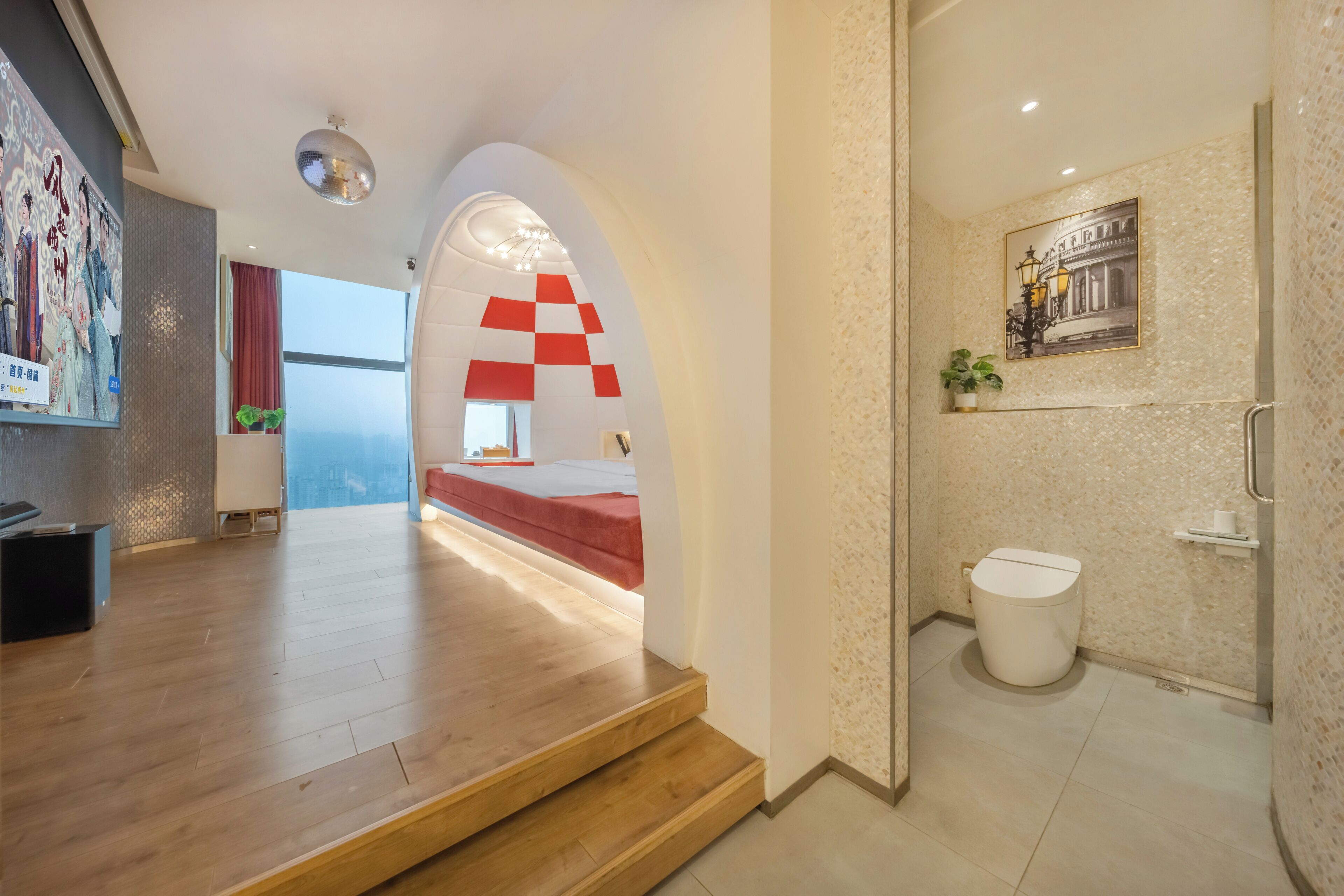 Une chambre d’hôtel avec un plancher en bois, un lit recouvert d’une courtepointe rouge, une salle de bain avec des toilettes et un lavabo, et une grande fenêtre donnant sur un beau paysage.