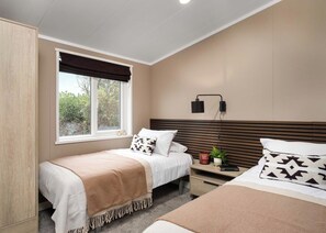 A bedroom with two beds, a window with a view, a bedside table with a lamp, and a wall-mounted light. - Premium modern and contemporary luxury lodge perfect for a relaxing escape. (Workington)