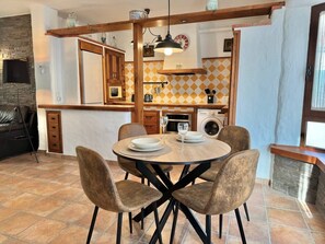 A dining area with a round table and four chairs, a kitchen in the background with a tiled backsplash, and a stone fireplace. - R007. Nueva 33 - Apartment N2 (Nerja)