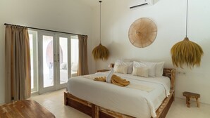 A bedroom with a wooden bed, a wooden nightstand, a small wooden stool, and a large fan on the wall. - Wellhouse Villas (Lembongan Island)