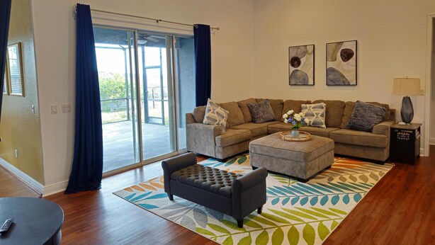 A spacious living room with a large sectional sofa, a small ottoman, and a coffee table. There is a sliding glass door leading to a balcony, and the room has hardwood flooring. - Countryside Comfort 5 MIN to FAST / WEC & 15 min HITS 1 acre 2 KNG BED Near I-75 (Ocala)