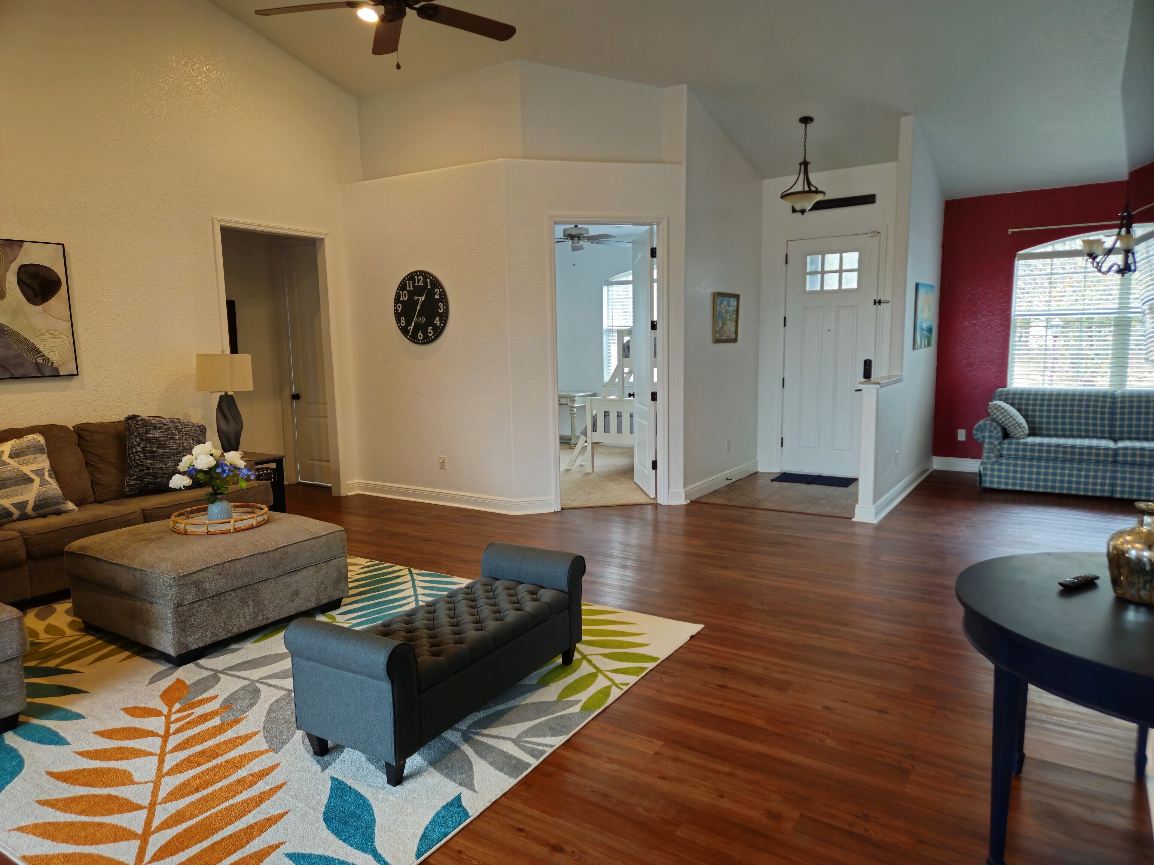 A spacious living room with hardwood flooring, a grey sofa, a black ottoman, and a round coffee table. A colorful rug with leaf patterns is on the floor.