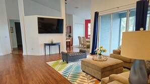A spacious living room with hardwood flooring, a large flat-screen TV, a grey sofa, a beige sofa, and a coffee table with a vase of flowers. - Countryside Comfort 5 MIN to FAST / WEC & 15 min HITS 1 acre 2 KNG BED Near I-75 (Ocala)
