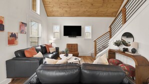 Una sala de estar moderna con techo de madera, un sofá de cuero negro, un sillón marrón, una mesa de centro blanca y una escalera con barandilla de madera.