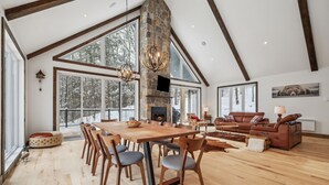 Ein geräumiges Wohnzimmer mit großem Holzesstisch, bequemen Sitzgelegenheiten, einem Steinkamin und großen Fenstern mit Blick auf schneebedeckte Bäume.