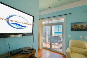 A room with a TV displaying 'Ocean Wave Rentals,' a balcony with a view of the ocean, a chair, and a painting. - 3BR Luxury Vacation Home - Unbeatable Location and View (Myrtle Beach)