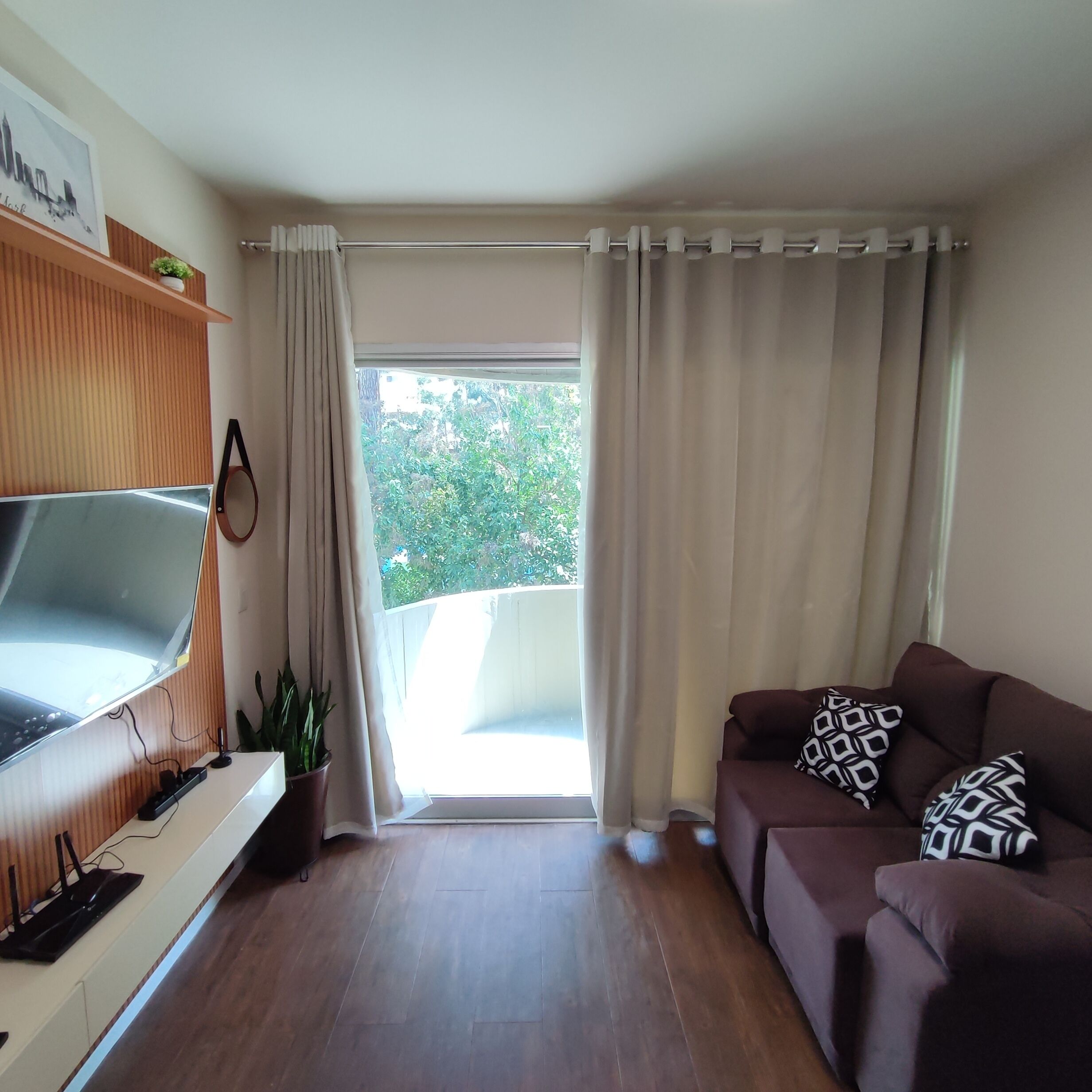 A living room with a flat-screen TV, wooden panel wall, potted plants, a brown sofa with patterned cushions, and a large window with curtains.