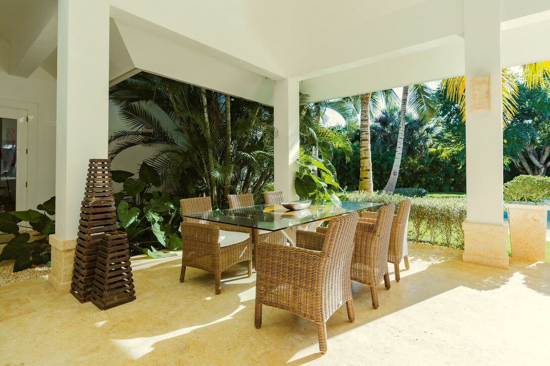 A covered outdoor dining area with wicker furniture and a glass table, surrounded by lush tropical plants.