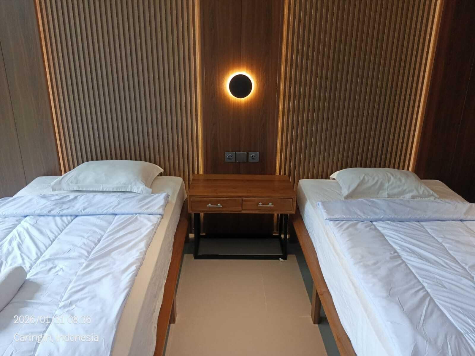 A hotel room with two beds, a wooden nightstand with a circular light, and a wooden panel wall.