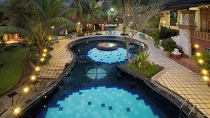 A pool area with a circular design, surrounded by palm trees and a building with a tiled roof. - The Village Resort Bogor Powered by Archipelago (Caringin)