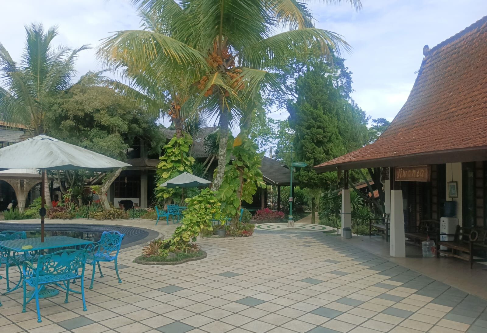 Un patio con suelo de baldosas, mobiliario exterior con sillas y sombrillas azules, palmeras y un edificio con tejado de tejas.