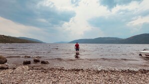 A person wading in shallow water near a rocky shore with a boat in the background.