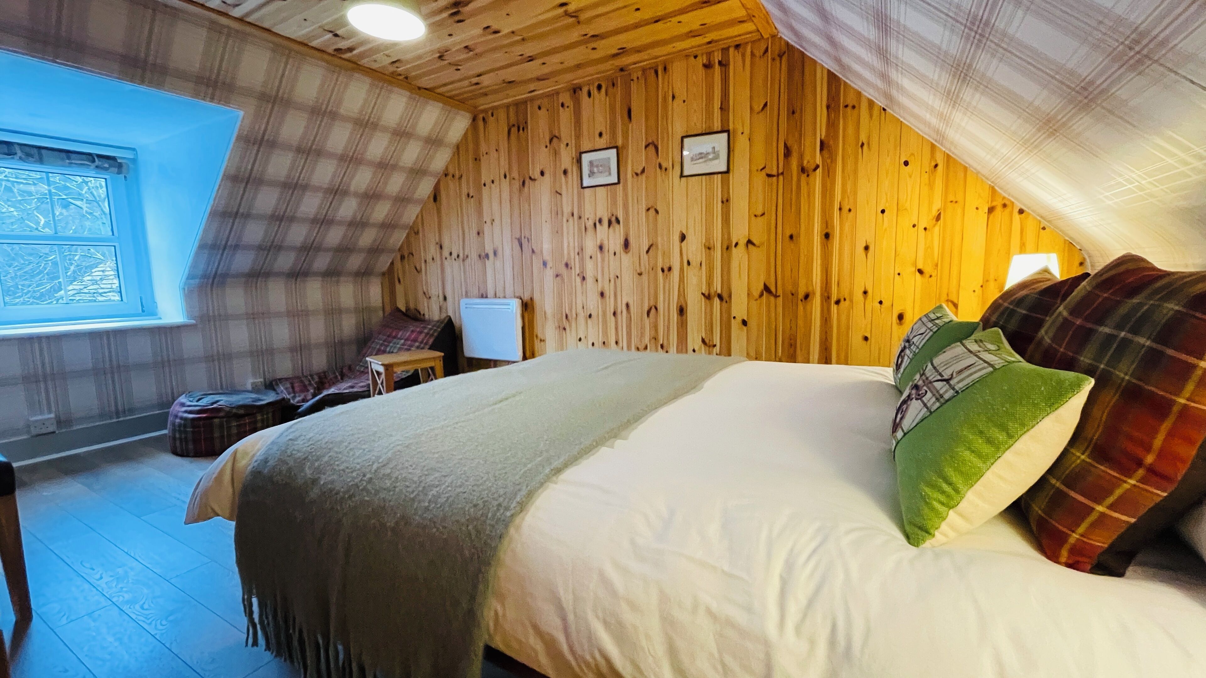 A cozy bedroom with a wooden ceiling, a bed with plaid pillows, a small table, and a window with a view.