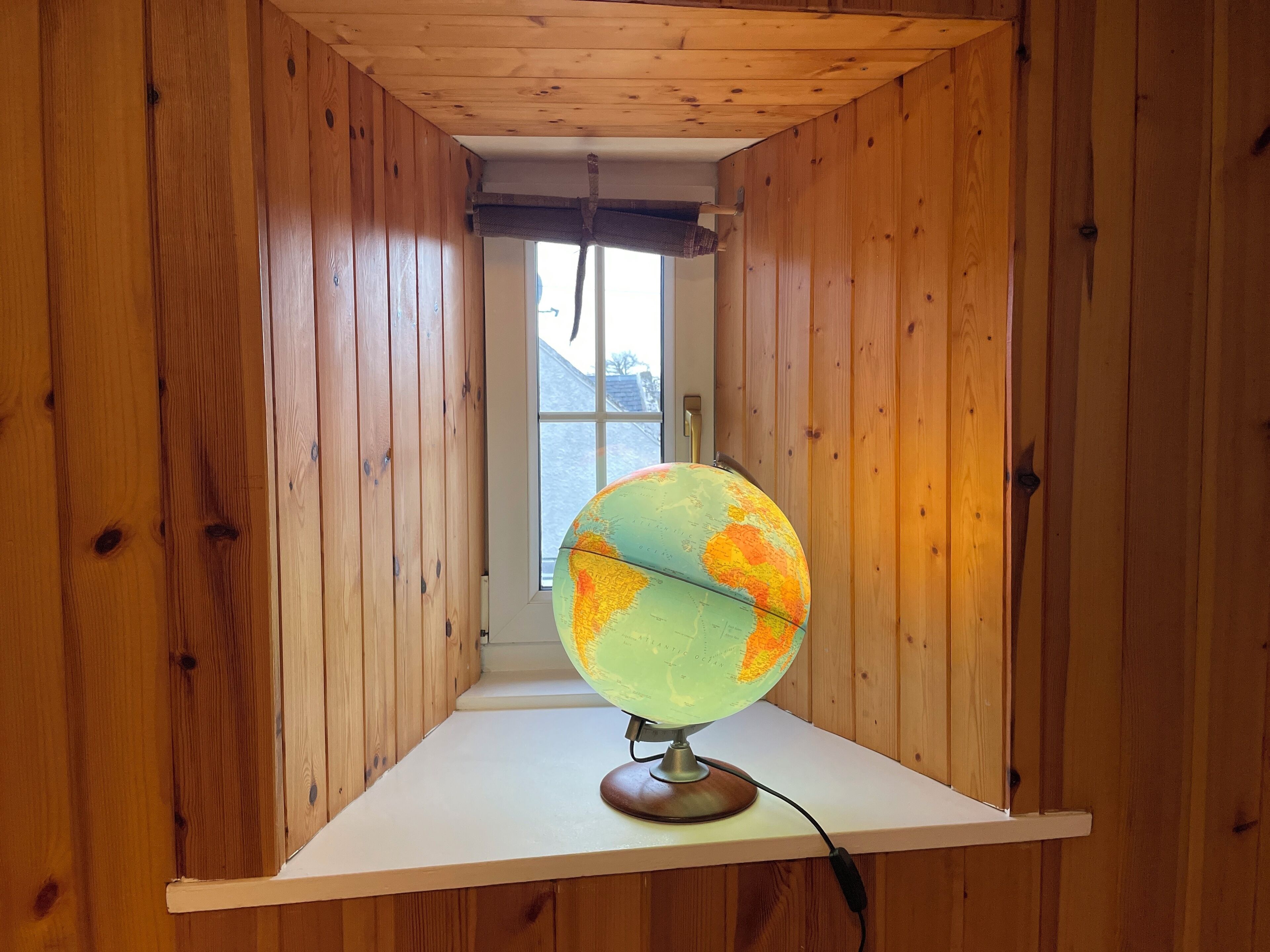 A globe on a wooden shelf in a room with wooden walls and a window.