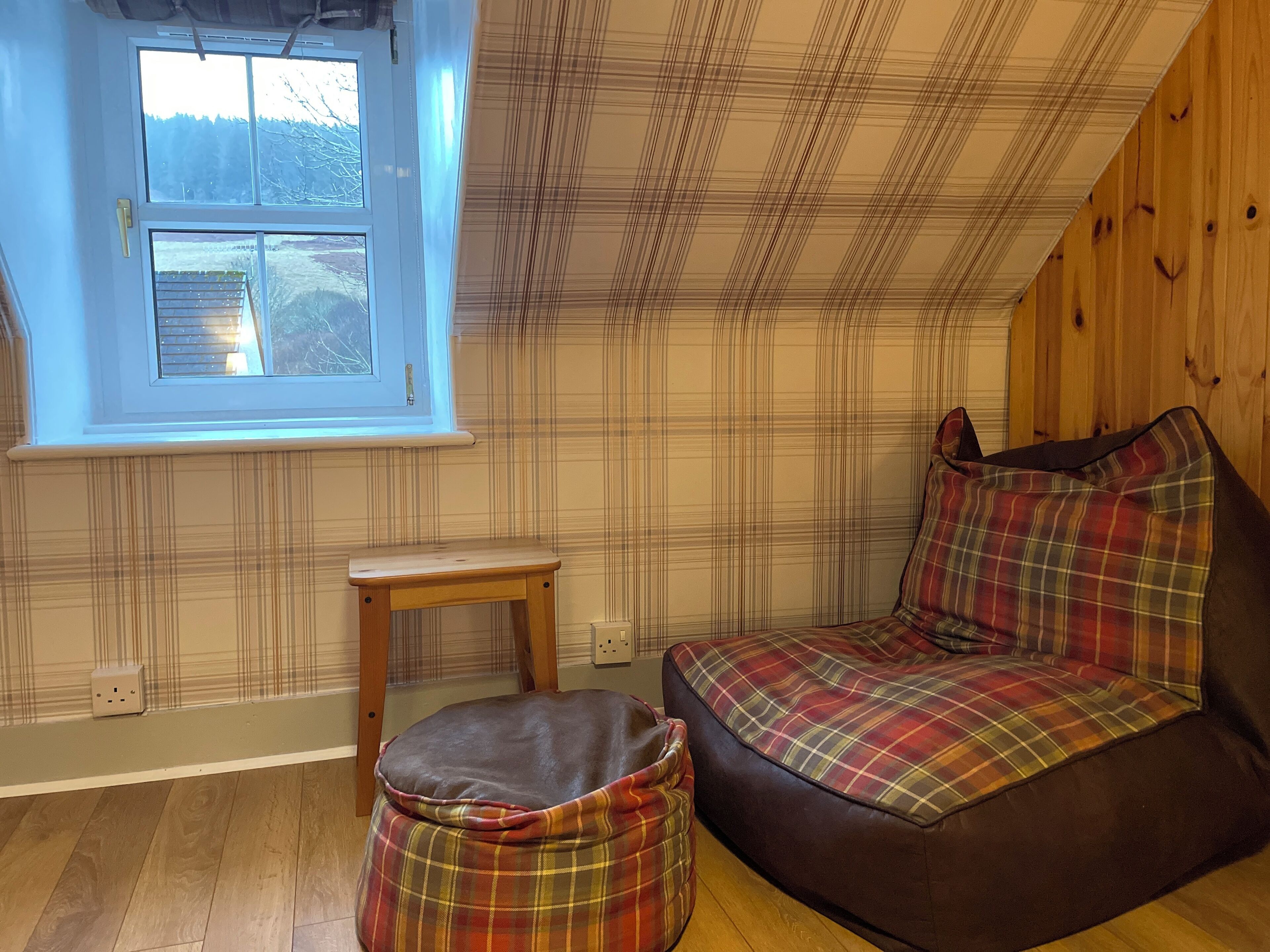 A cozy room with a plaid-patterned sofa, a small wooden table, and a plaid ottoman. A window with blinds is visible, and the room has wooden walls and flooring.