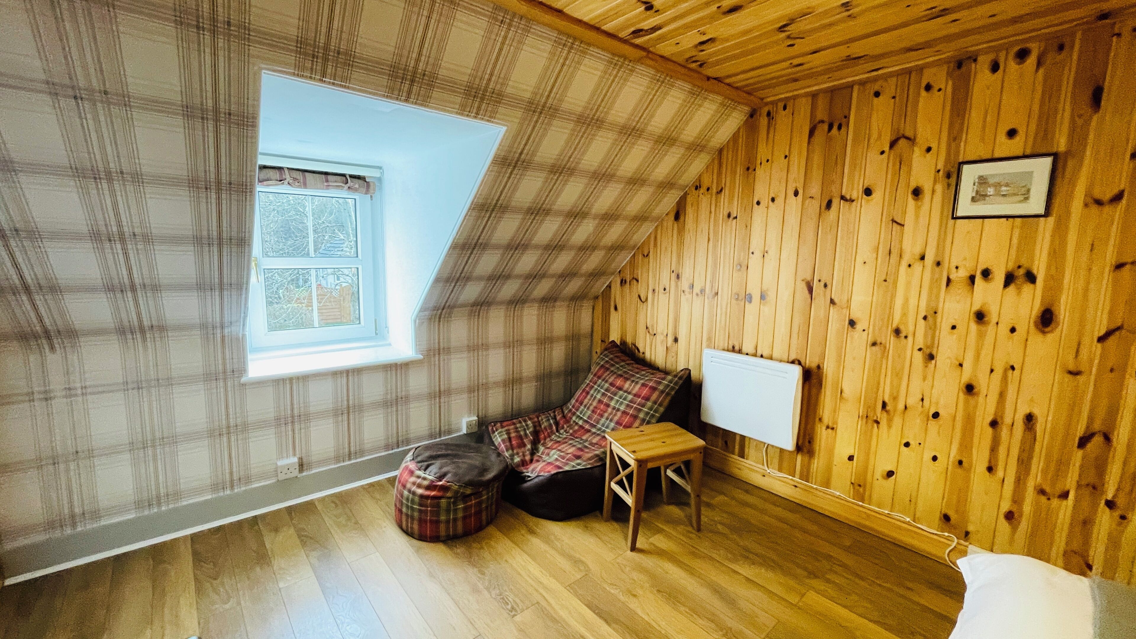 Une chambre mansardée chaleureuse, avec un plafond en pente, une fenêtre ornée de rideaux à carreaux, un pouf à carreaux, une petite table et un radiateur blanc.