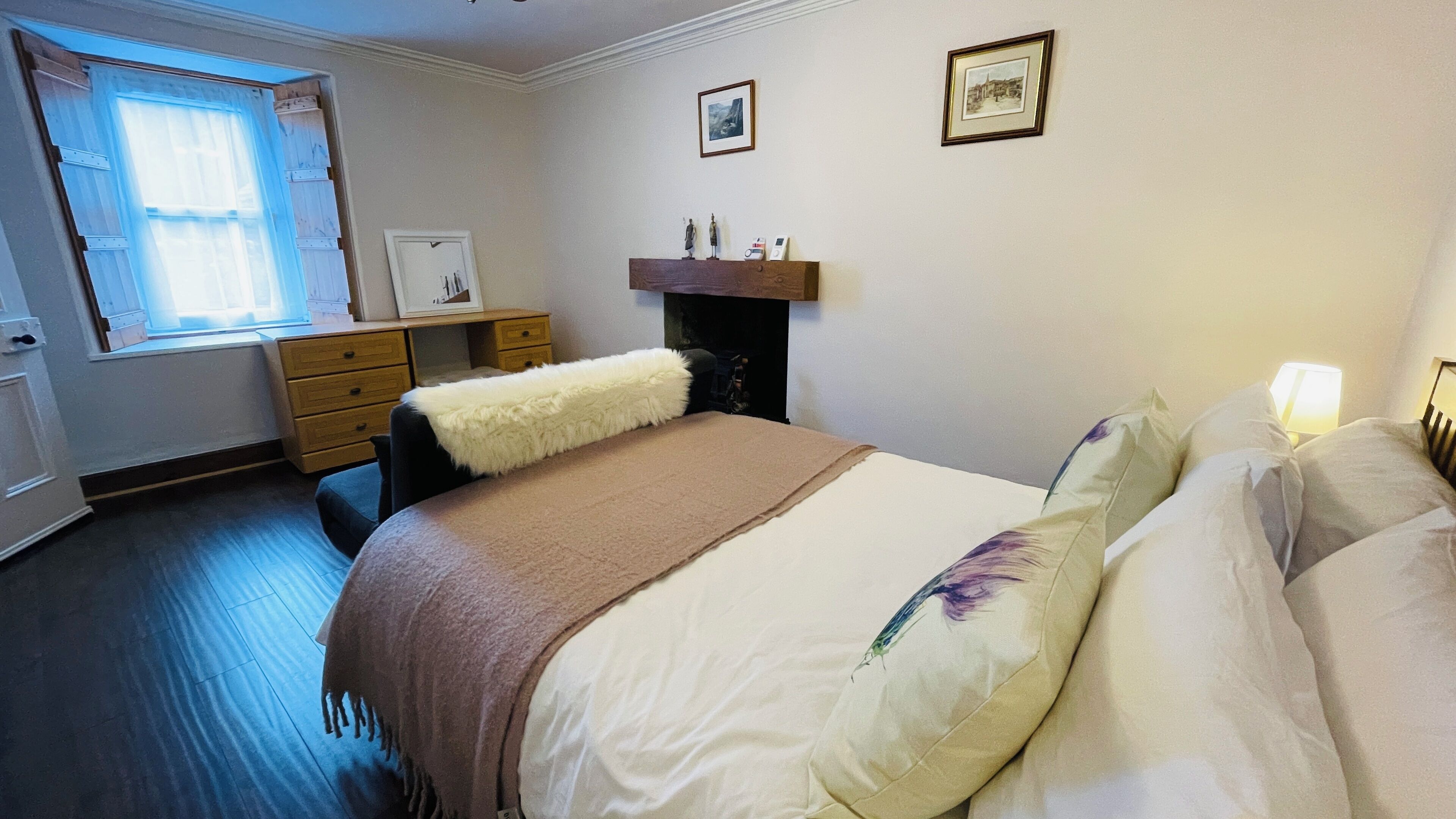 Une chambre à coucher comprenant un lit, une commode, une fenêtre avec des rideaux, une cheminée et deux tableaux encadrés accrochés au mur.