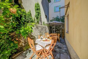 A stone patio with a table set for dining, surrounded by greenery and a building with a window.