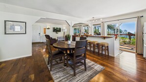 A spacious dining area with a wooden table and chairs, a kitchen in the background, and a view of the outdoors through large windows. - Black Sands Escape- Ocean and Range Views- Comfort (Whitethorn)