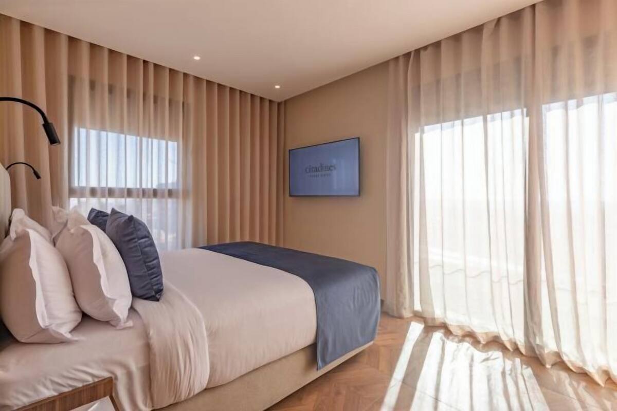 Una habitación de hotel moderna con cama, cortinas, un televisor montado y suelo de madera.