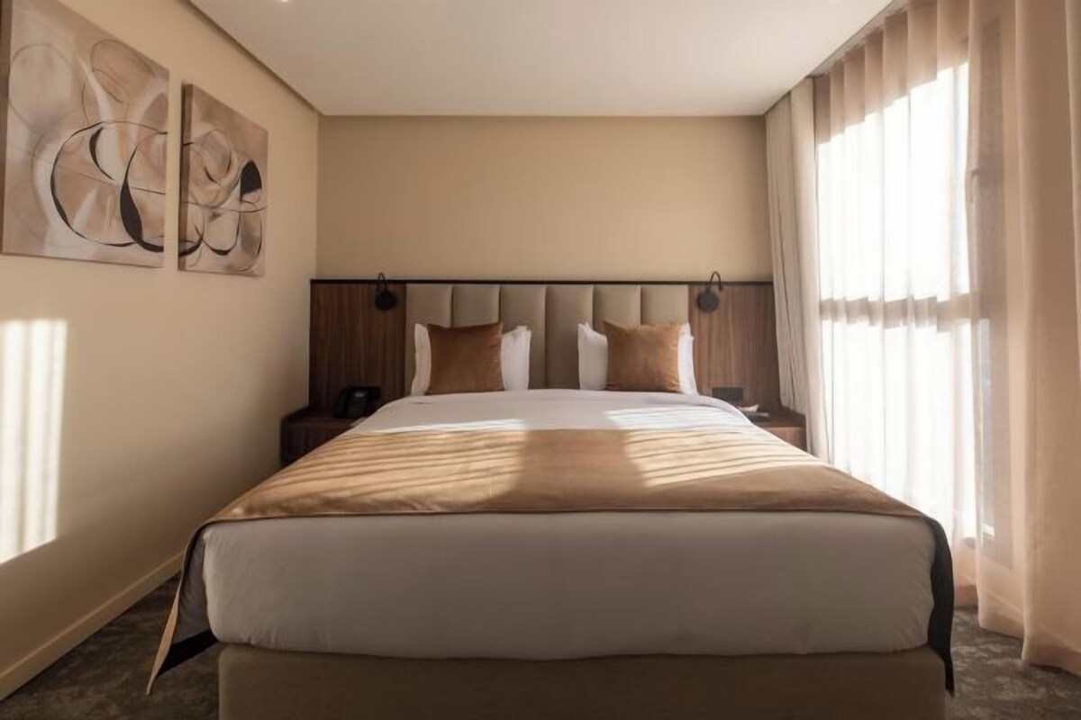 Ein Hotelzimmer mit einem großen Bett, einem Kopfteil, einem Nachttisch, einer Wandleuchte und einem Fenster mit Vorhängen.