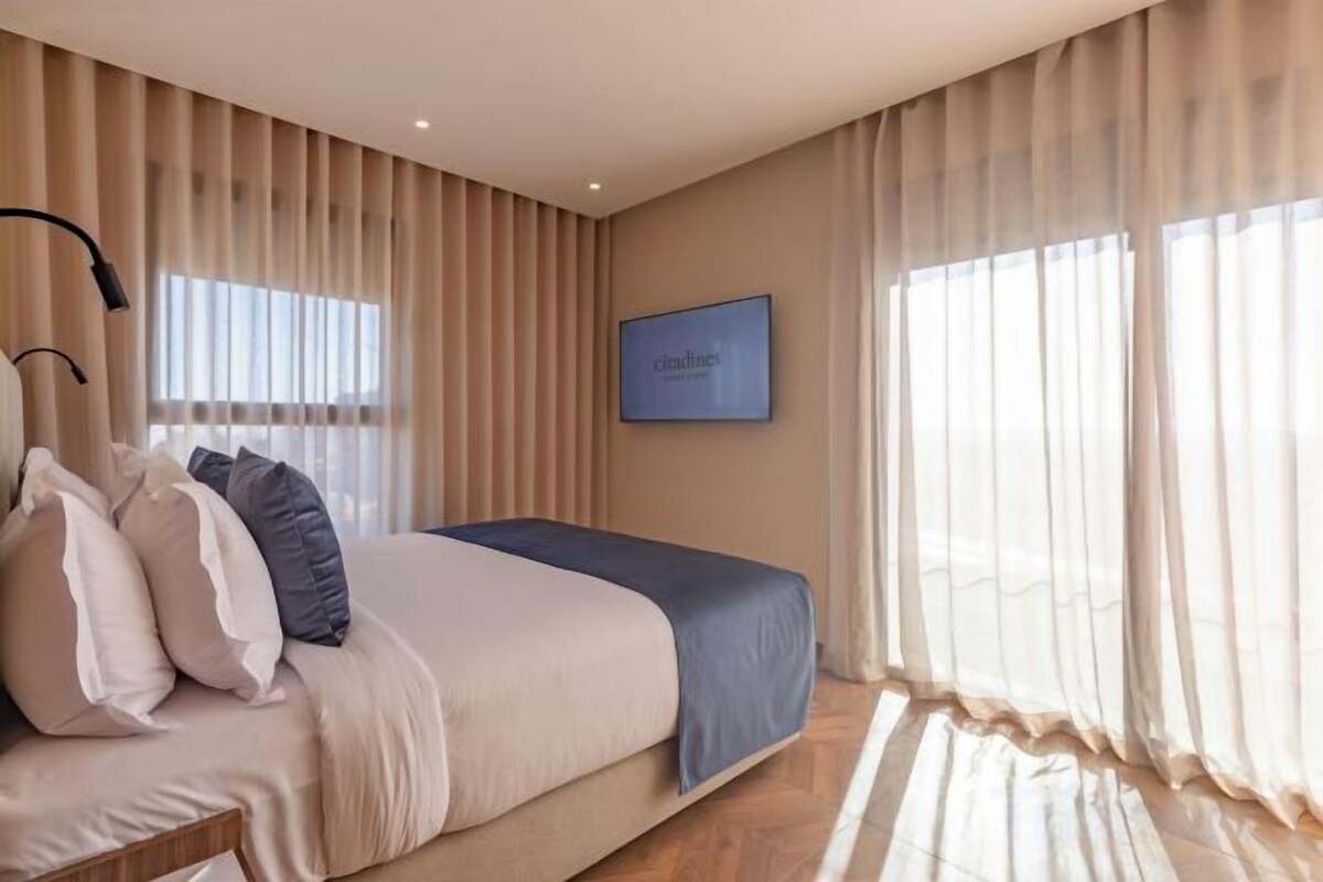 Quarto com cama, cortinas, TV na parede e janela.