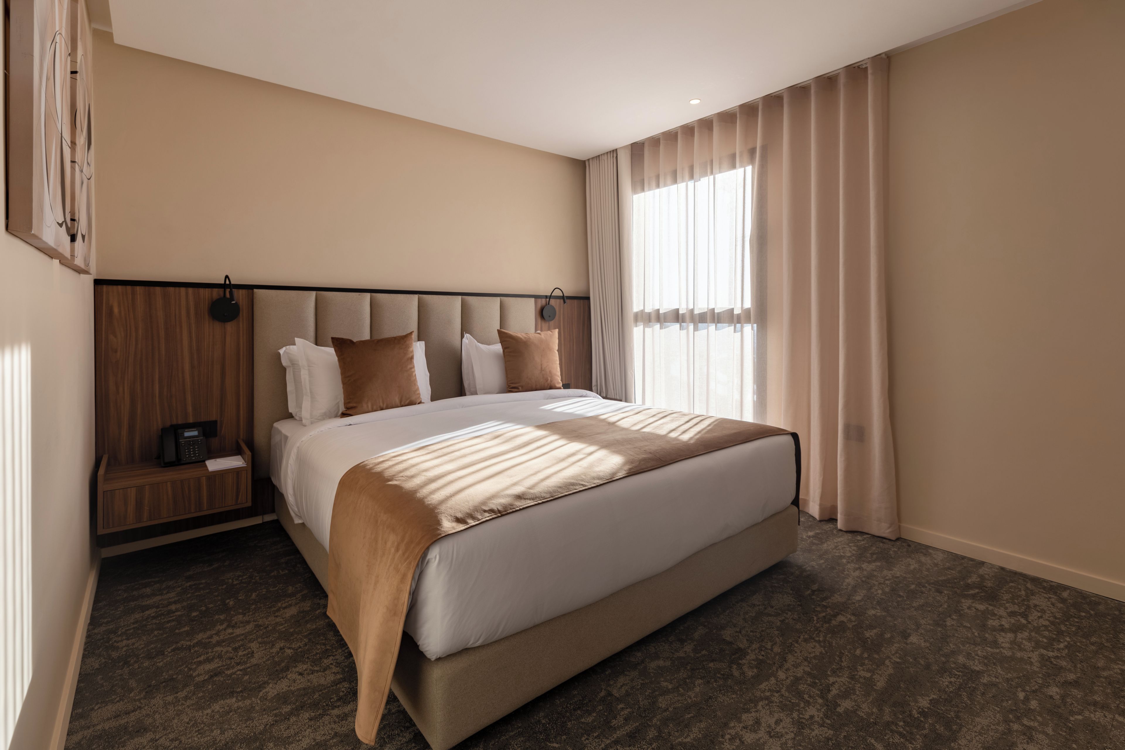 Une chambre d’hôtel moderne avec un grand lit, une table de chevet en bois, un téléphone et une fenêtre avec des rideaux.