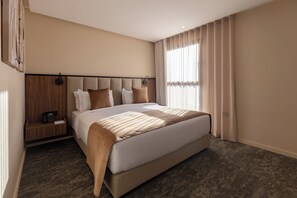Una habitación de hotel moderna con una cama grande, una mesita de noche de madera, un teléfono y una ventana con cortinas.