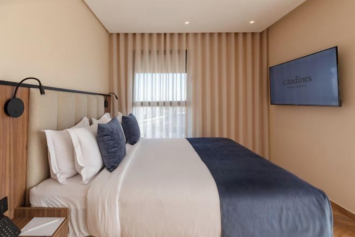 Une chambre d’hôtel moderne dotée d’un grand lit, d’un téléviseur à écran plat et d’une fenêtre avec des stores verticaux.