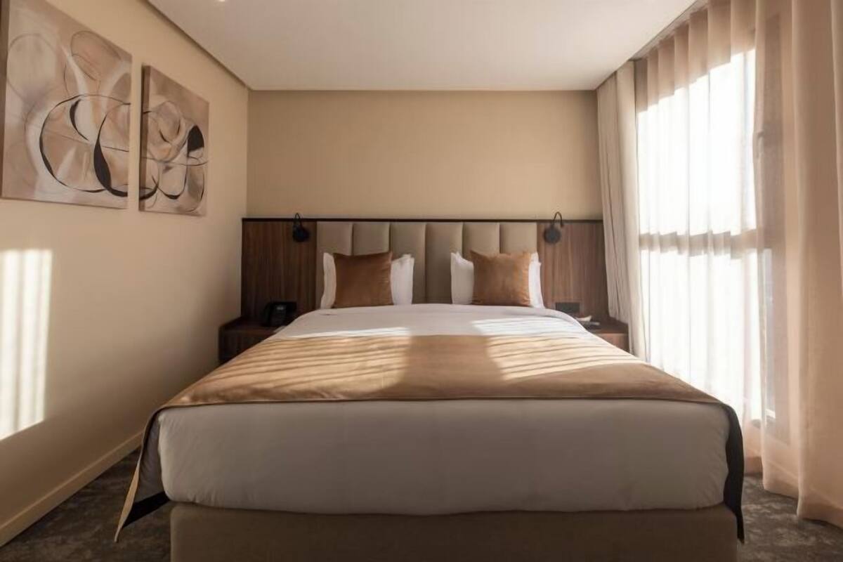 Una habitación de hotel con una cama grande, cabecera, mesita de noche, lámpara fijada a la pared y una ventana con cortinas.