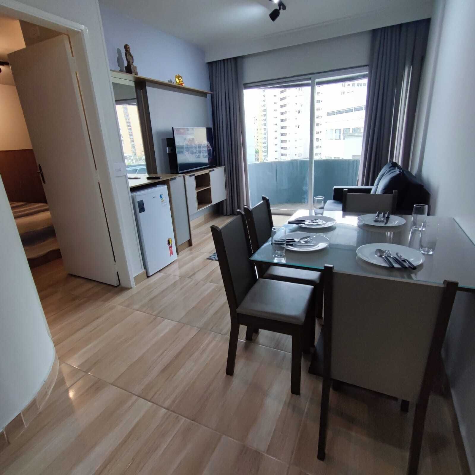 A modern kitchen with a dining area, a small refrigerator, and a television on a shelf.