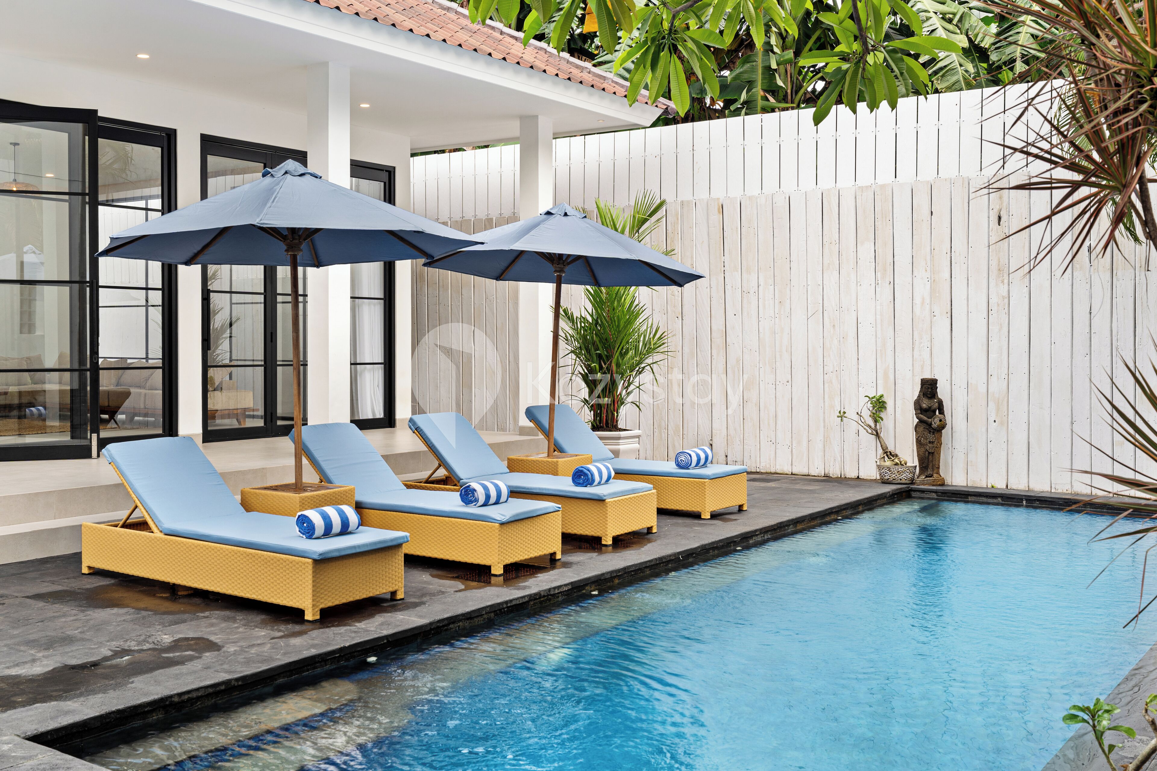 Uma área à beira da piscina com espreguiçadeiras e guarda-sóis, um cercado branco e uma estátua.